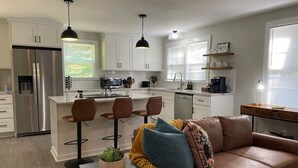 Una cocina moderna con armarios blancos, un refrigerador de acero inoxidable y una isla central con taburetes. Hay un sofá de cuero marrón con cojines y una mesita con una planta.