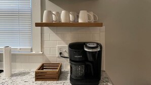 Una cocina con cafetera, un cesto de madera y una estantería con cristalería y tazas.