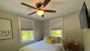 Un dormitorio con cama, ventilador de techo, dos ventanas con persianas, una mesita de noche con lámpara y una cita enmarcada en la pared.