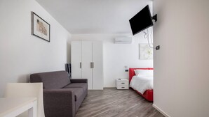 Ein modernes Zimmer mit grauem Sofa, weißem Schreibtisch, rotem Bett und einem Wand-Fernseher.