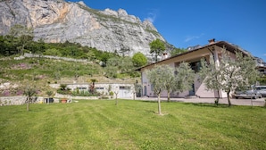 A house with a mountain backdrop, a grassy yard, and a car parked nearby. - Studio 'Monte Brento Gaggiolo 2' with Mountain View and Private Garden (DRO)
