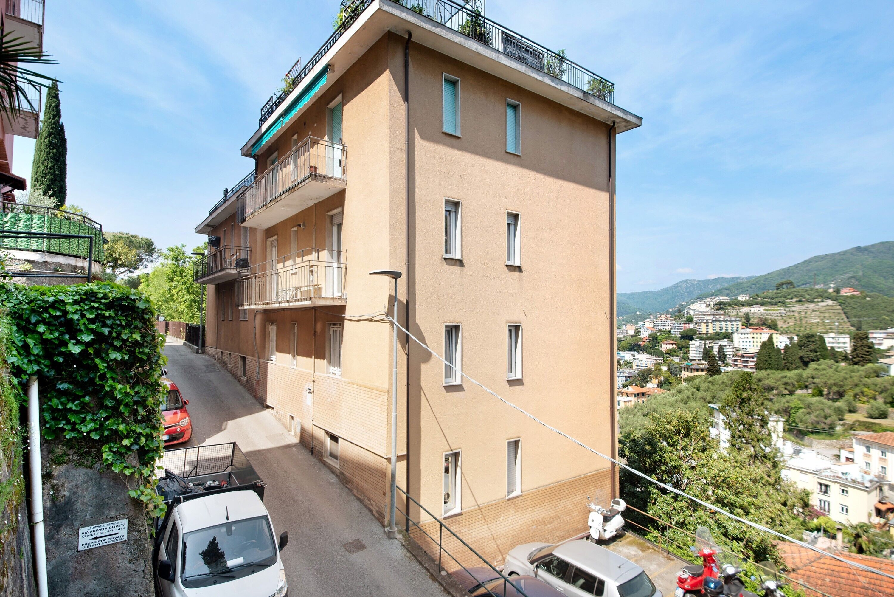A multi-story building with balconies, a narrow street, parked cars, and a view of a town with hills in the background.