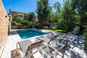 Un espace aménagé au bord de la piscine, avec des chaises longues et une table, entouré de verdure et d’un mur en pierre.