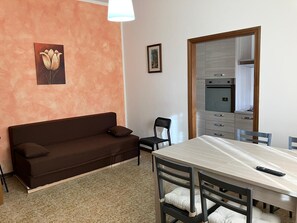 A room with a sofa, a dining table with chairs, a kitchen area, and a framed picture on the wall.