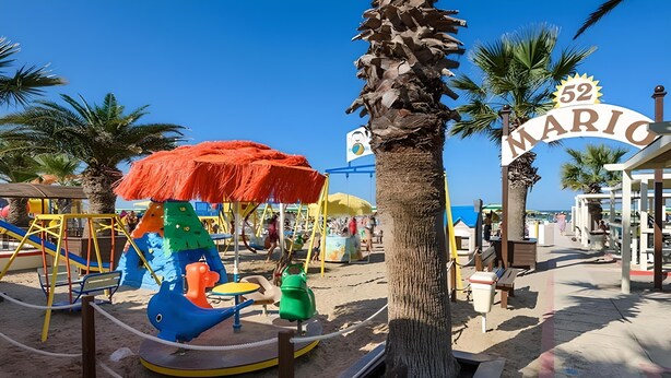 Zona de playa con área infantil, que incluye un carrusel y columpios, y un letrero que dice '52 MARIO'.