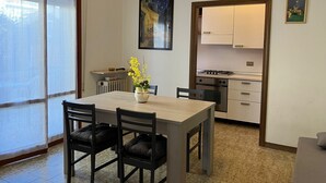 Un comedor con una mesa y sillas, un florero con flores, un cuadro en la pared y una cocina visible a través de una puerta abierta.