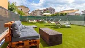 Una terraza en la azotea con un sofá de madera, sillones de descanso y un otoman de mimbre sobre césped artificial.