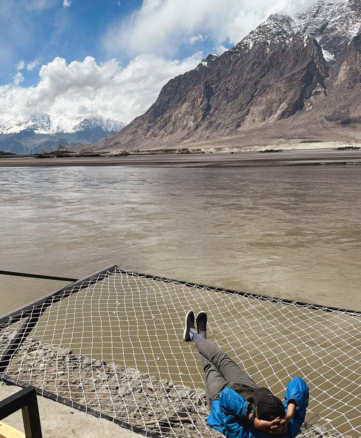A person lying on a netted platform by a river with mountains in the background.