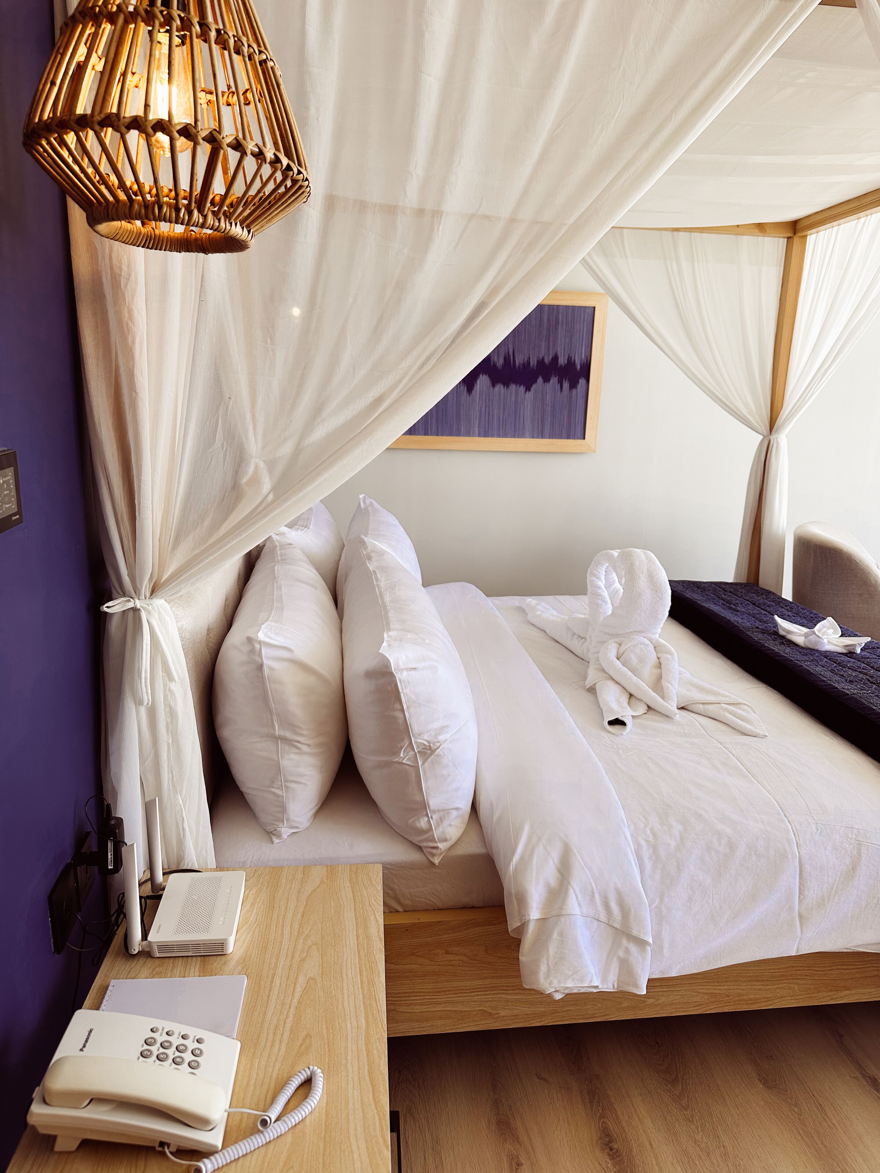 A bedroom with a canopy bed, white bedding, a bedside telephone, and a projector on a wooden desk.