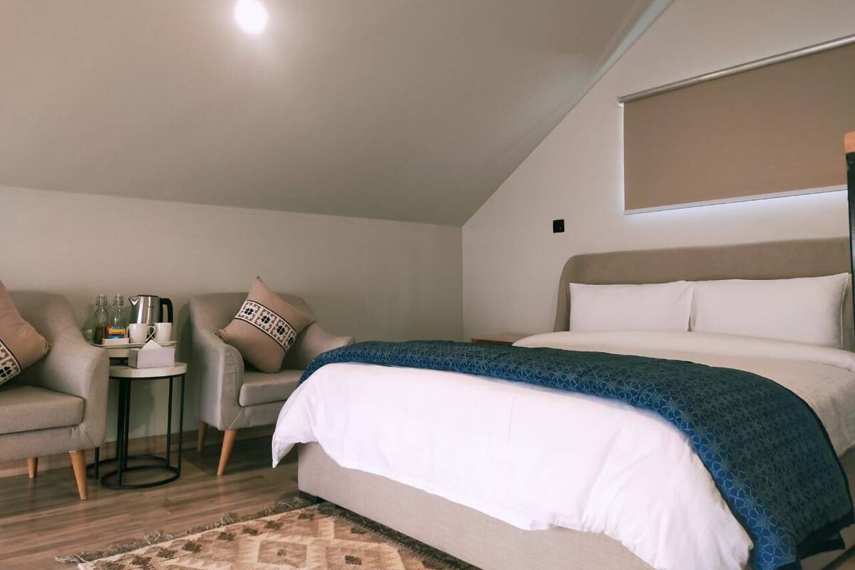 Ein Schlafzimmer mit einem Bett, zwei Sesseln, einem kleinen Tisch mit Gegenständen und einem Fenster mit Jalousien.