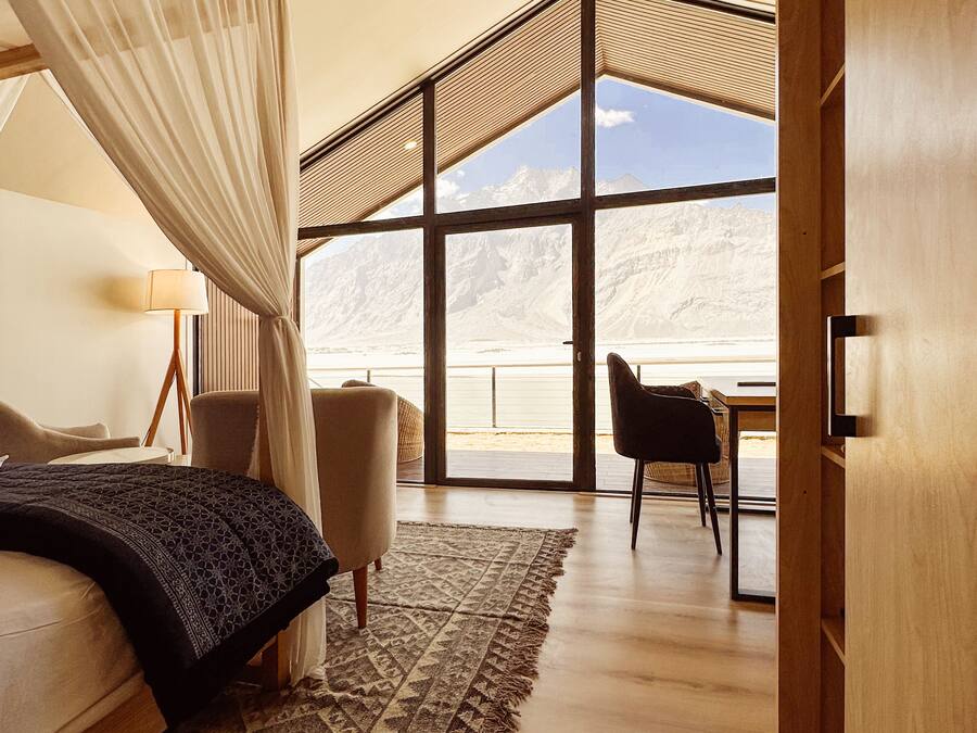 A bedroom with a large bed, a chair, a desk, and a view of mountains through large windows.