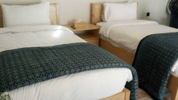 A room with two beds, each with a wooden headboard, white linens, and a blue patterned blanket. There is a wooden nightstand with a tissue box and a remote control.