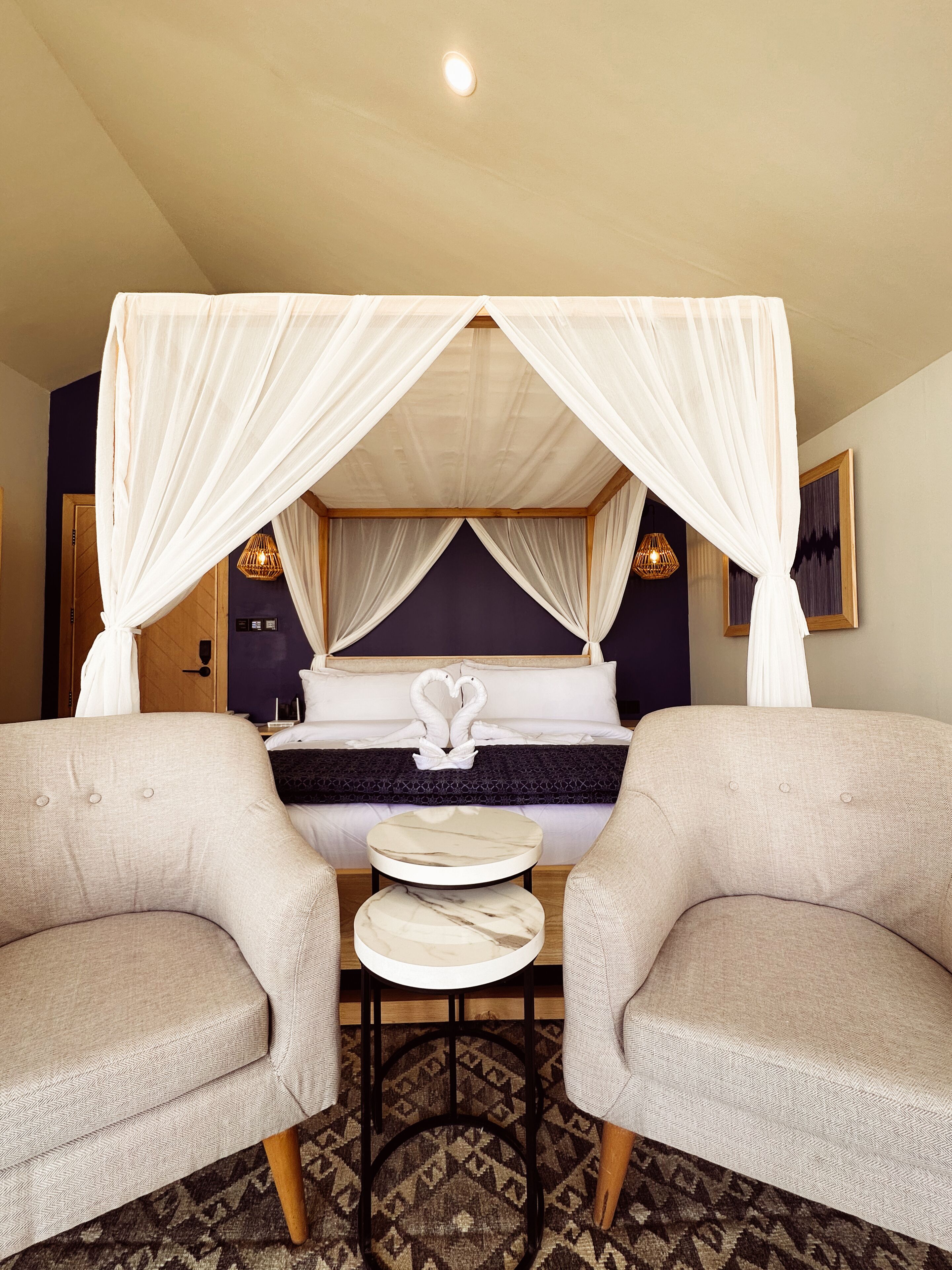A bedroom with a canopy bed, two armchairs, a small round table, and a patterned rug.