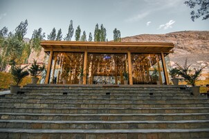 A wooden pavilion with a glass enclosure, surrounded by stone steps and potted plants, set against a mountainous backdrop.