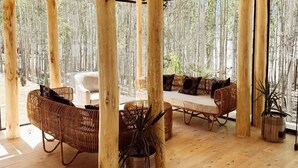 Interior de una cabaña de madera con techo alto, ventanas amplias y una zona de estar acogedora con muebles de mimbre y plantas en macetas.
