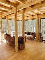 A wooden cabin interior with a high ceiling, large windows, and a cozy seating area with wicker furniture and potted plants.