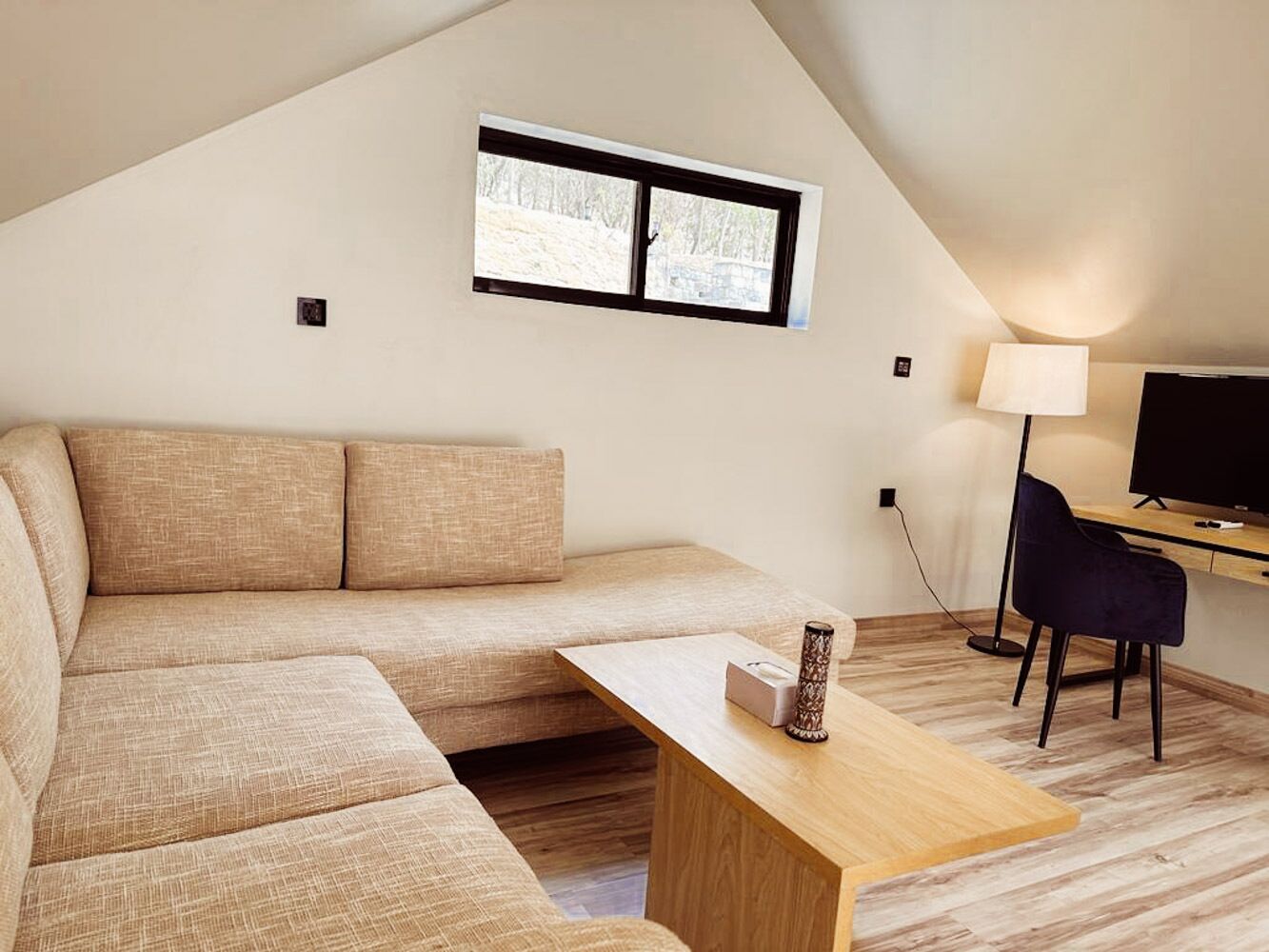 A modern, well-lit room with a sofa, a wooden table, a desk with a chair, and a TV.