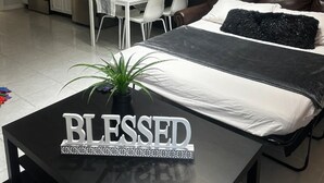 Ein modernes Schlafzimmer mit Bett, Nachttisch mit einem Schild, einer Essecke mit Tisch und Stühlen sowie einer Küche im Hintergrund.