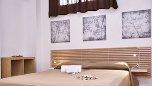 Un lit bien fait, recouvert d’une couvre-lit beige, avec deux oreillers et un petit tas de coquillages. Trois œuvres d’art sont accrochées au mur et représentent des branches et des feuilles.