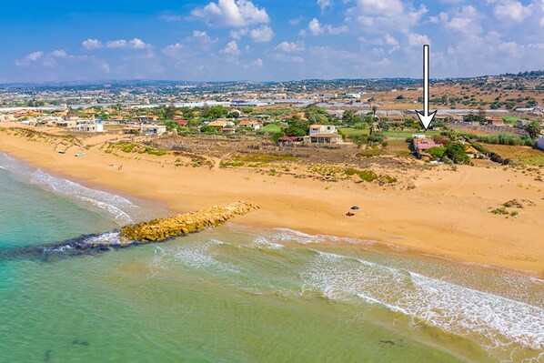 A coastal area with a sandy beach, a rocky breakwater, and scattered buildings.
