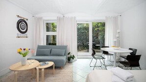 Ein moderner, gut beleuchteter Raum mit einem grauen Sofa, einem kleinen runden Tisch mit Blumenvase, einem Esstisch mit Stühlen und einem großen Fenster mit Blick auf Grünfläche.