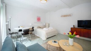 Ein modernes Hotelzimmer mit Bett, Esstisch, einer Couch, einem Fernseher und einem Fenster mit Vorhängen.