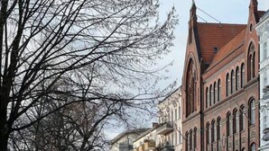 Eine Kopfsteinpflasterstraße, gesäumt von mehrstöckigen Gebäuden, mit parkenden Autos und einem gelben Lieferwagen.