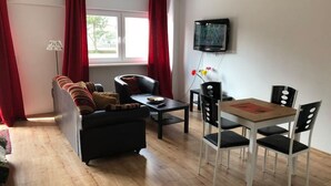 Een woonkamer met een bank, een salontafel en een eettafel met stoelen. Er hangt een televisie aan de muur.
