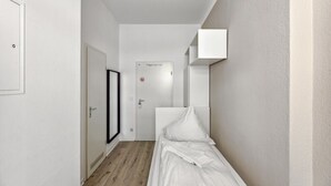 A narrow hallway with a bed on the right, a door with a no entry sign, and a white cabinet on the wall.