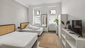 A modern hotel room with two single beds, a TV, a mirror, and a window with blinds.