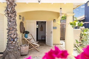 Entrada de uma casa de praia, com uma palmeira, uma placa indicando 'BEACH HOUSE' e uma porta de entrada.