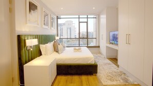 A modern bedroom with a green upholstered headboard, a large bed, a television, and a view of a cityscape through a large window. - Azalia Apartments (Sandton)