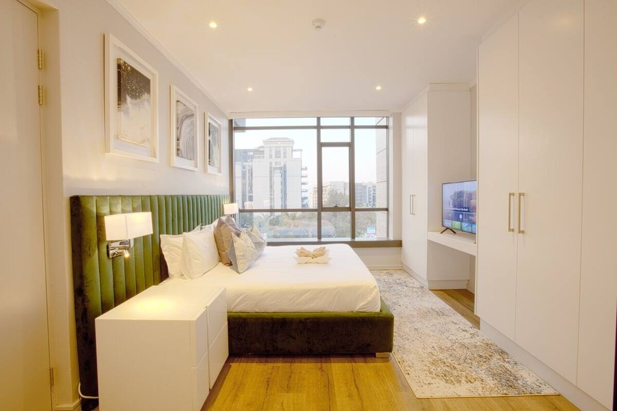 A modern bedroom with a green upholstered headboard, a large bed, a television, and a view of a cityscape through a large window.
