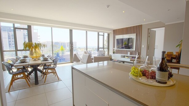A modern kitchen with a large island, dining area with chairs, and a view of the city through large windows. - Azalia Apartments (Sandton)
