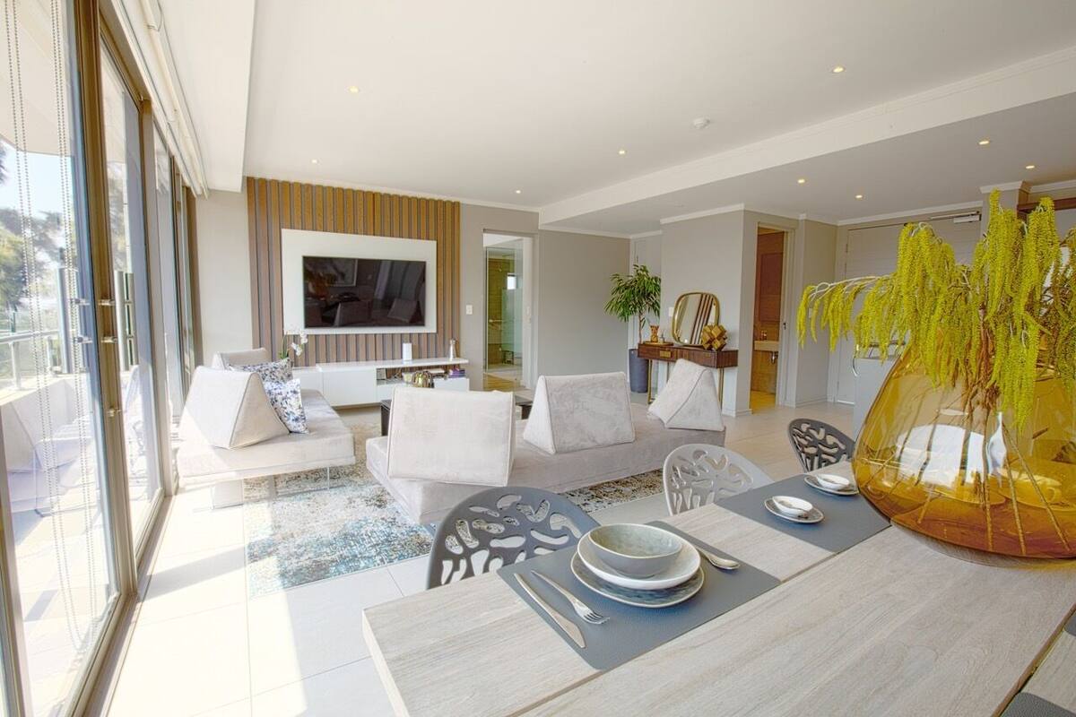 A modern living room with a sofa set, a dining table with plates and cutlery, and a large window.