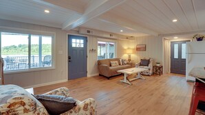 A spacious living room with a wooden floor, a sofa, a coffee table, and a view of the outdoors through large windows. - Big Pine Island Lake Cottage w/ Boat Dock & Kayaks (Belding)