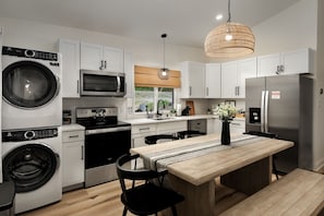 Una cocina moderna con armarios blancos, un refrigerador de acero inoxidable, una lavadora y un comedor con mesa y sillas de madera.