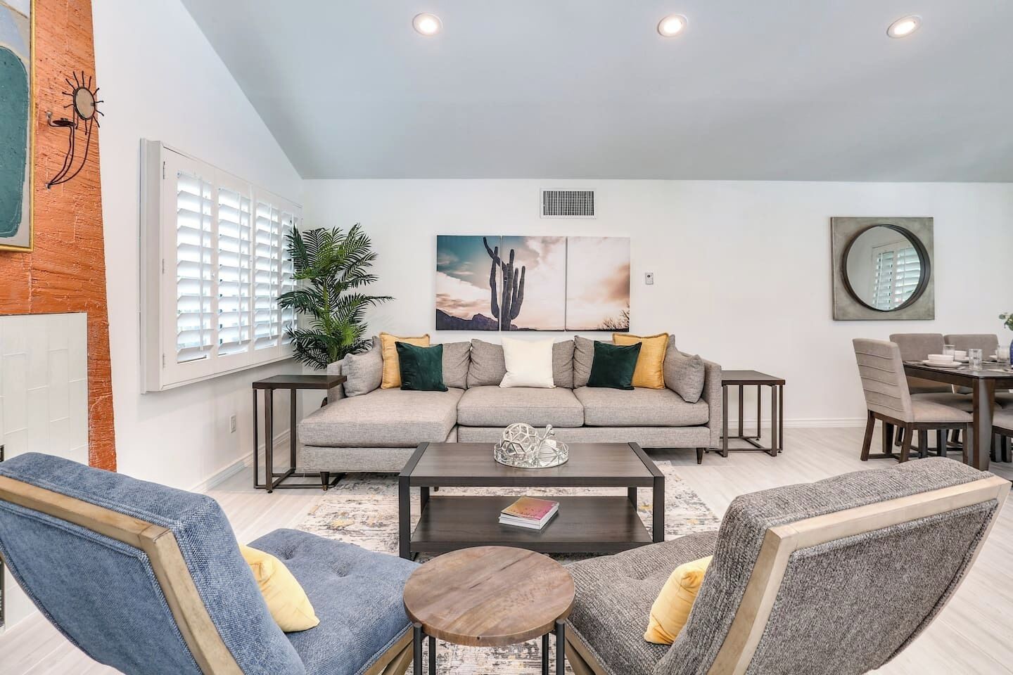 A living room with a sofa, coffee table, and chairs, a dining area with a table and chairs, and a large window with blinds.