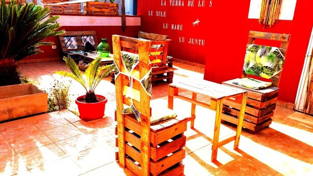 A patio area with wooden furniture, potted plants, and a red wall with text. - Tobia's House  (Giuggianello)