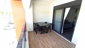 A balcony with a wooden floor, a table with chairs, and a sliding glass door leading to a dark interior. - Çamlık Apart (Ayvalik)