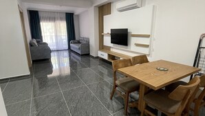 A modern living room with a flat-screen TV, a wooden dining table with chairs, a sofa, and a large window with curtains. - Çamlık Apart (Ayvalik)