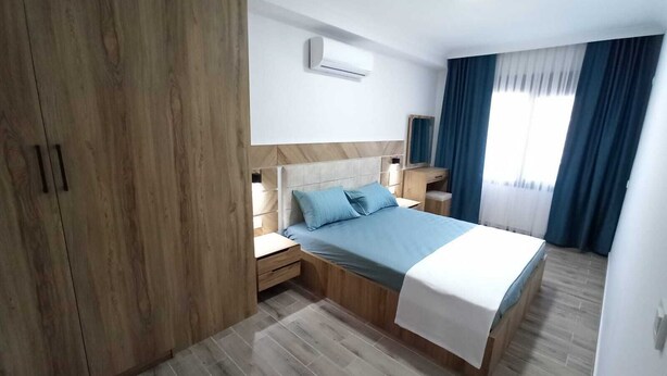 A modern bedroom with a wooden wardrobe, a bed with blue bedding, a bedside table, an air conditioning unit, and a window with curtains. - Çamlık Apart (Ayvalik)
