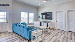 A spacious living room with a blue sofa, a wooden coffee table, a TV mounted on the wall, and a ceiling fan. - Get ready to enjoy Sunny Daze on the coast (Rockport)