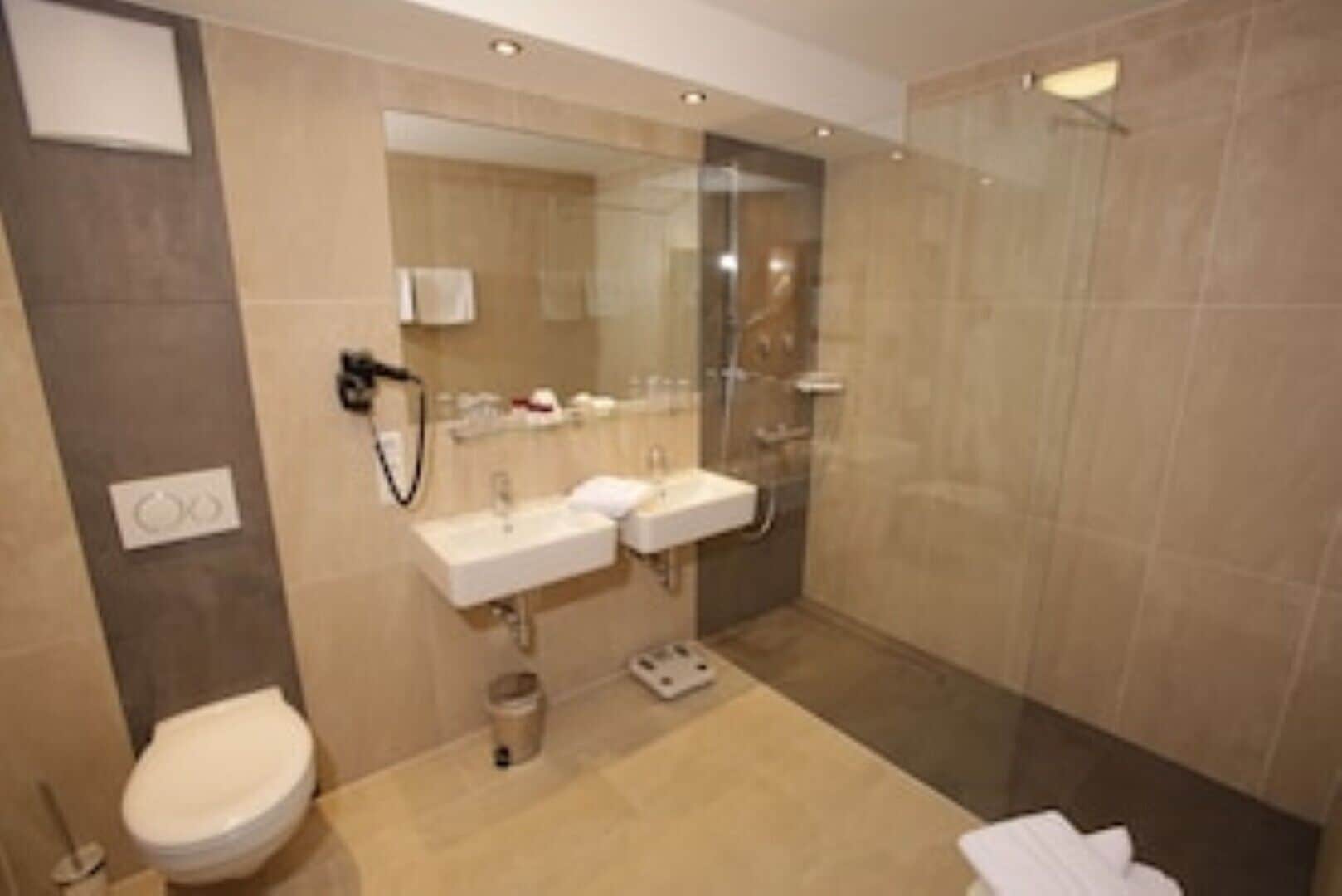 Une salle de bain moderne équipée de toilettes, de deux lavabos, d’une douche à parois de verre et d’un porte-serviettes.