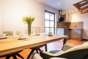 A dining area with a wooden table, chairs, a vase with yellow flowers, and a kitchen in the background.