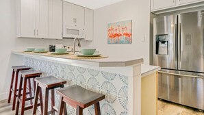 A modern kitchen with white cabinets, a stainless steel refrigerator, a microwave, and a bar area with stools. - Sea Street - Holiday stays at the beach! (St. Augustine Beach)