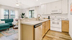 A modern kitchen with white cabinets, a dishwasher, an oven, and a microwave. There is a green sofa and a small dining table with a plant. - Sea Street - Holiday stays at the beach! (St. Augustine Beach)
