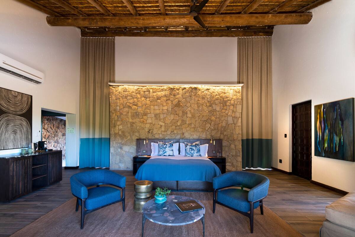 A modern bedroom with a stone accent wall, a large bed, two blue armchairs, a coffee table, and a TV on a wooden cabinet.