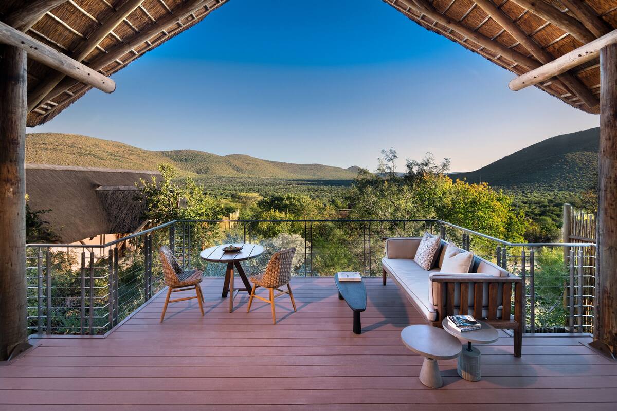 A wooden deck with a thatched roof, a round table, two wicker chairs, a sofa, and a small table with books.