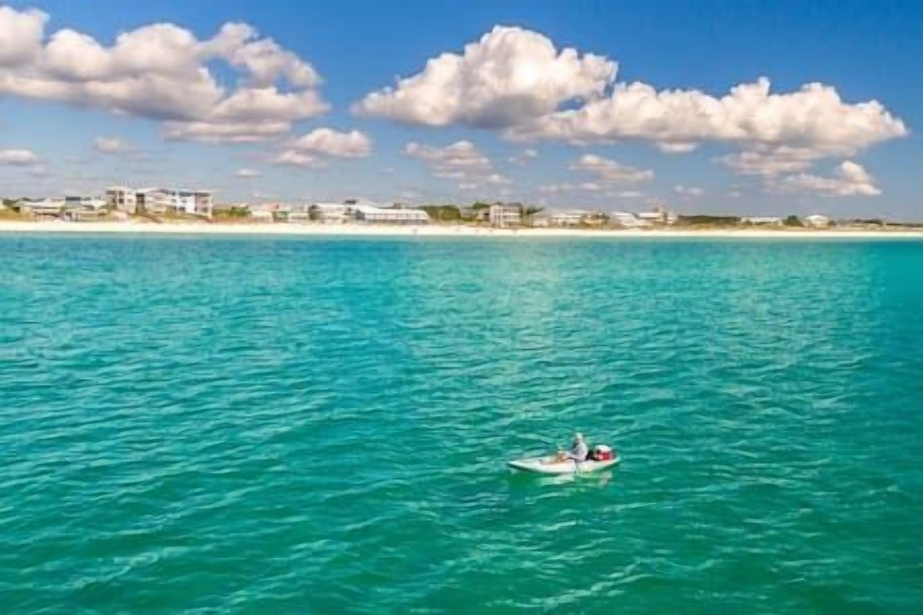 A coastal scene with a clear blue sea, a small boat with two people, and a sandy beach with buildings in the background.